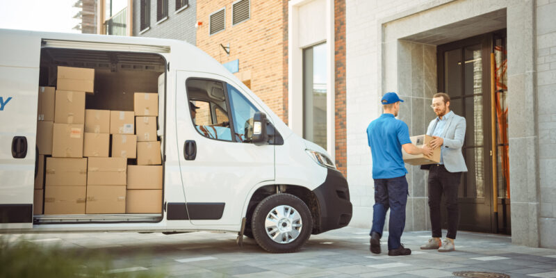 Delivery Man Gives Postal Package to a Business Customer, Who Signs Electronic Signature POD Device. In Stylish Modern Urban Office Area Courier Delivers Cardboard Box Parcel to a Man